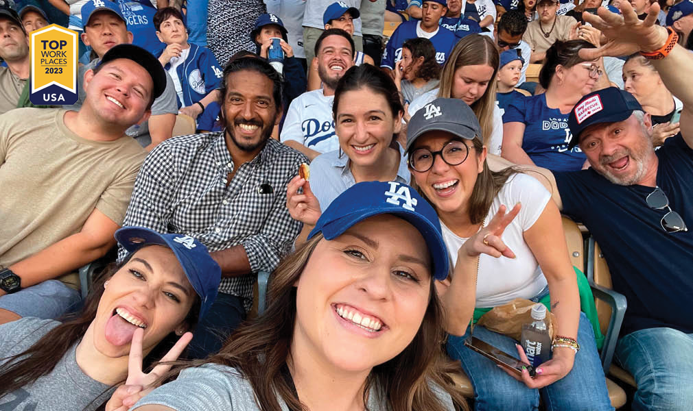 MGAC Awards Top Workplaces. An MGAC team are at a baseball game. They are wearing blue hats, and sit crowded together, smiling and making silly faces at the camera.