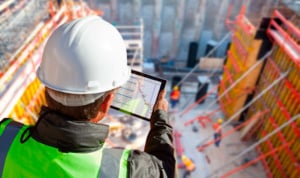 A worker looks over a construction jobsite, holding a digital device that shows construction plans.