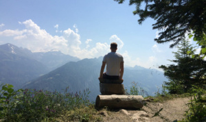 MGAC Impacts: Alex Pearce sits on a log, facing away from the camera and overlooking a mountain range.